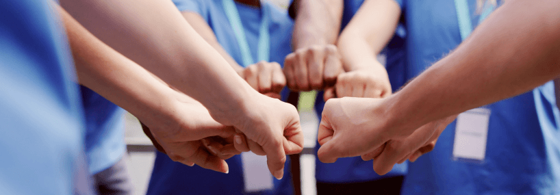 A group of people in blue shirts join their fists together in a circle, symbolizing teamwork and unity.